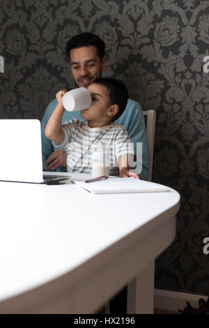 Jeune père est assis à la table avec son fils sur ses genoux. Son fils est en train de copier ce qu'il a vu son père en train de faire et a ses pères chope à sa bouche, Banque D'Images