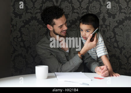 Jeune père de famille est en train de rire comme des copies de son fils lui. Il est de porter le téléphone à l'oreille de ses fils, qui fait semblant de parler à quelqu'un. Banque D'Images