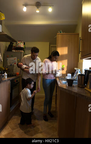 Jeune famille dans la cuisine. La mère est lave-vaisselle pendant que le père est maintenant leur bébé et de tendre à son aîné qui a blessé son doigt Banque D'Images