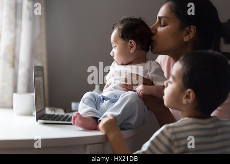 Jeune mère assise avec ses deux fils à la table à manger. Elles sont regarder les dessins animés sur un ordinateur portable et la mère embrasse son bébé sur la tête. Banque D'Images