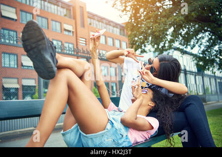 Femme tendue sur banc avec ami prendre une photo à l'extérieur par de grands arbres dans une grande clôture Banque D'Images