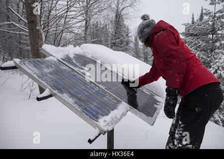 Une dame dans une veste rouge efface la neige de l'panneaux solaires après une tempête dans la région de Hastings Highlands, Ontario, Canada. Banque D'Images