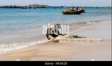 Pêcheur assis sur une plage de sable blanc privée dans le soleil du matin fixant son filet avec ses mains pour prendre plus de poisson, au Vietnam. Banque D'Images