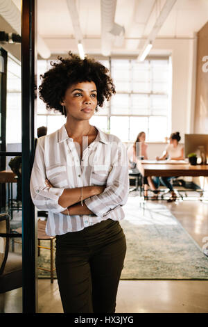 Portrait of young African woman leaning à office de porte et à la voiture. Femme sérieuse à l'exécutif office avec des personnes travaillant en arrière-plan. Banque D'Images