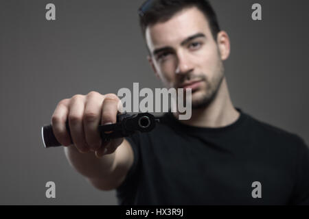 Criminel dangereux qui menace des armes à feu incliné holding visant à huis clos. Portrait de l'atmosphère contrastée gris sur fond de studio. Banque D'Images