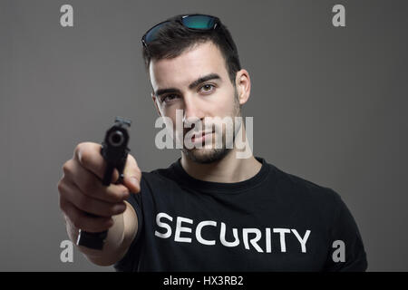 Agent de sécurité pensée holding gun dans une main visant à vous. Portrait de l'atmosphère contrastée gris sur fond de studio. Banque D'Images