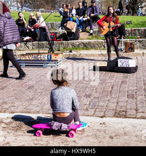 Mauer Park, Berlin, Allemagne, le 26 mars 2017. Les horloges déplacé vers l'avant aujourd'hui et se sont aventurés à l'extérieur pour profiter des Berlinois le chaud soleil du printemps dans les parcs de la ville. Un enfant assis sur un skate-board jouit de la musique d'un artiste de rue.Eden Breitz/Alamy Live News Banque D'Images