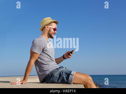 Portrait d'un côté heureux jeune homme assis au bord de la mer avec un téléphone mobile Banque D'Images