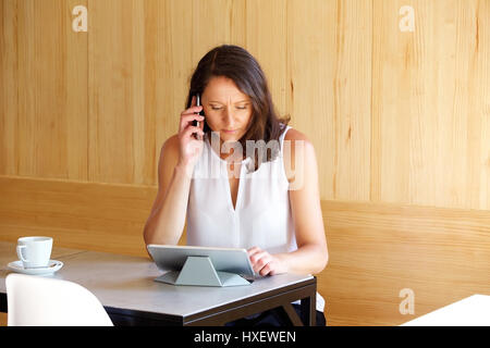 Jolie femme plus âgée dans un café, talking on mobile phone et recherche sur ordinateur Tablet Banque D'Images