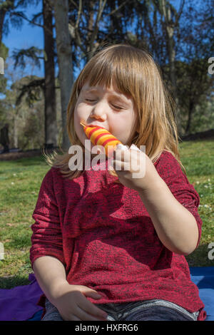 Portrait de trois ans, bénéficiant de l'alimentation de l'enfant blond orange colorés et red ice lolly ou glace, assis sur l'herbe verte champ dans parc public R Banque D'Images