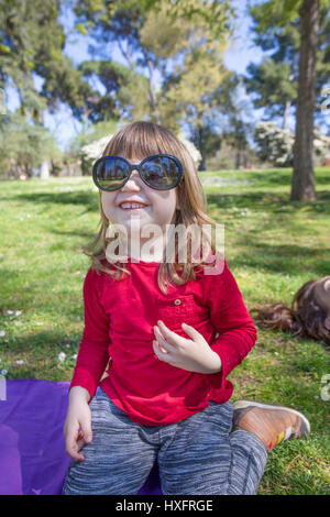 Funny portrait of blond trois ans enfant avec chemise rouge assis dans l'herbe verte dans le parc, à côté de sa mère, jouant avec des femme adulte noir Banque D'Images