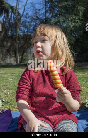 Portrait de trois ans, l'enfant blonde holding orange colorés et red ice lolly ou glace, assis dans une serviette bleue sur l'herbe verte dans les champs Banque D'Images