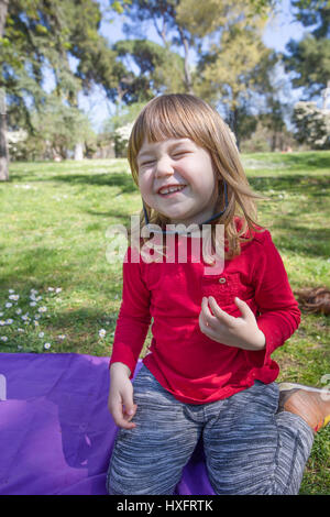 Funny portrait of blond trois ans de l'enfant, avec chemise rouge assis dans l'herbe verte en parc, rire et jouer avec les femmes adultes su surdimensionnée noire Banque D'Images