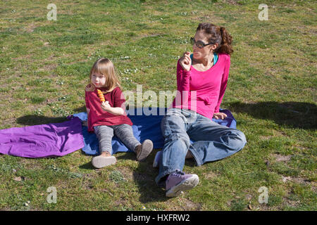 Femme mère, avec des jeans et chemise rose, et trois ans de l'enfant blond eating ice lolly, ou glace, assis dans une serviette bleue sur l'herbe verte fie Banque D'Images
