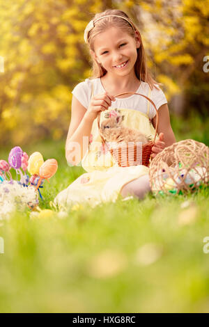 Beautiful smiling little girl holding cute bunny dans le panier et assis sur l'herbe avec des oeufs de Pâques en vacances de printemps. En regardant la caméra. S COPIE Banque D'Images