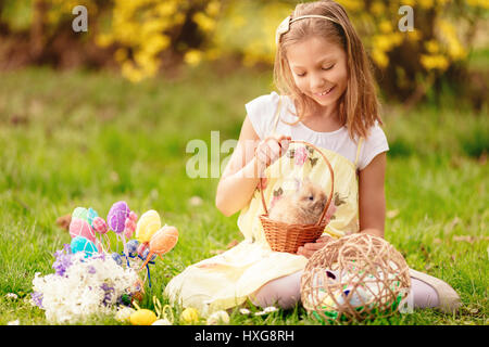 Beautiful smiling little girl holding cute bunny dans le panier et assis sur l'herbe avec des oeufs de Pâques en vacances de printemps. Banque D'Images