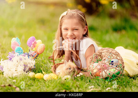 Beautiful smiling little Girl with cute bunny dans le panier et oeufs de Pâques sur l'herbe en vacances de printemps. En regardant la caméra. Banque D'Images