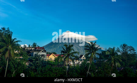 Vue lointaine du volcan Mayon, Legazpi, Philippines avec de petits nuages de maisons et palmiers dans l'avant-terrain. Banque D'Images