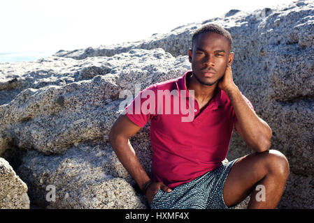 Portrait de jeune homme africain assis sur le rocher à la plage avec expression grave Banque D'Images