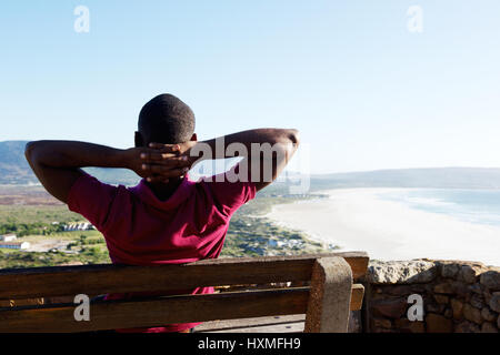 Vue arrière portrait de jeune homme africain assis détendu sur un banc avec ses mains derrière la tête, jeune homme en vacances. Banque D'Images