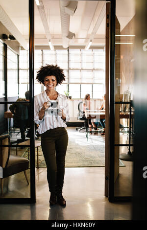 Full Length portrait of smiling young woman standing in office porte avec du café. Femme ayant pause café avec les personnes qui travaillent dans backgrou Banque D'Images