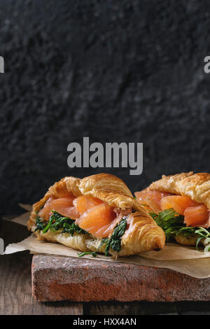 Deux croissants avec du saumon salé, fumé et à la roquette épinard servi sur du papier sulfurisé conseil terre cuite ancienne en bois foncé sur arrière-plan. Copy space Banque D'Images