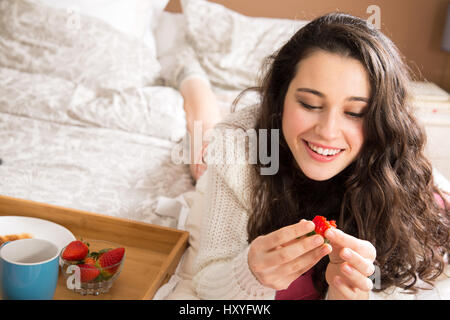 Belle jeune femme ayant le petit déjeuner au lit et manger des fraises Banque D'Images