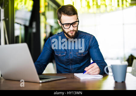 L'homme en prenant des notes en bas de son nouvel ordinateur portable à l'œuvre dans l'office du matin avec le café Banque D'Images
