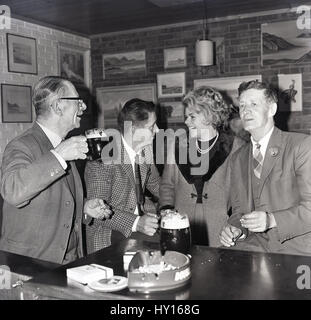 Années 1960, historiques, glamour femme dans un manteau avec un revers de fourrure dans un bar avec des collègues masculins de déguster une boisson et l'allume après le travail, England, UK Banque D'Images