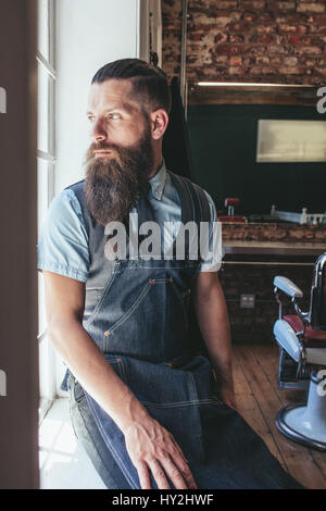 Coiffure homme sérieux avec longue barbe à l'extérieur de la fenêtre. Coiffure assis sur un rebord de fenêtre dans le salon et la pensée. Banque D'Images