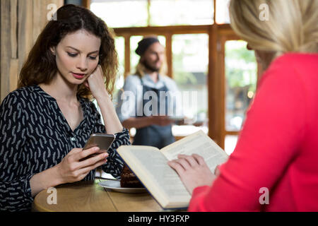 Les jeunes femmes à l'aide de Phone and reading book at table in coffee shop Banque D'Images