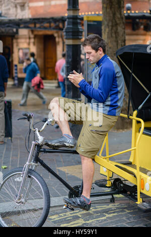 Jeune homme London pedicab driver (pilote de pousse-pousse) vérifie son téléphone mobile dans l'attente de son prochain billet. Young man using smartphone au travail, attendez Banque D'Images