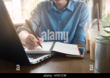 Young Asian woman writing on hipster portable en main droite tout en travaillant avec un ordinateur portable dans un café. Offres d'activité starup conc Banque D'Images