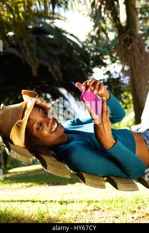 Side portrait of a smiling young woman relaxing on hammock with mobile phone Banque D'Images