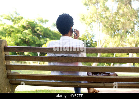 Portrait d'arrière young African woman sitting on a park bench using cell phone Banque D'Images