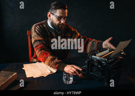 Poète barbu lunettes en tapant sur machine à écrire rétro. Livres sur le bureau avec une nappe bleu foncé vintage Banque D'Images