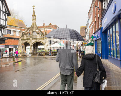 Couple de personnes âgées à l'aide d'un parapluie sur les rangs du Blue Boar près de la Croix de la volaille monument ancien, Salisbury, Wiltshire, Royaume-Uni. Météo mixtes dans la célèbre cathédrale avec de fortes averses de pluie d'avril et le soleil sur un après-midi de printemps. Banque D'Images