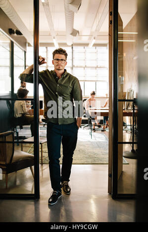 Portrait de jeune homme debout dans la porte de bureau de démarrage avec des gens qui travaillent en arrière-plan. Banque D'Images