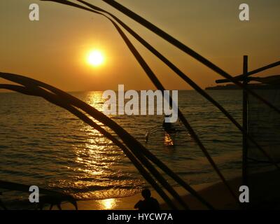 Le coucher du soleil et d'ossature d'un bateau de pêche sur la mer avec un garçon et un chien jouant dans le sable au premier plan Banque D'Images