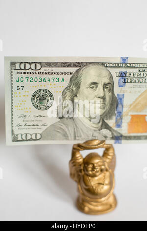 Une petite figurine bouddha doré debout sur un fond blanc est titulaire d'un one hundred dollar bill in United States service au-dessus de sa tête Banque D'Images