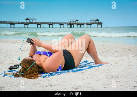 Grande taille jeune femme couchée sur plage de sable blanc de Clearwater Beach, FL à l'écoute de smart phone with earphones wearing bikini avec Pier 60 et le sh Banque D'Images
