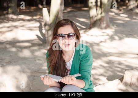 Belle jeune femme portant des lunettes de soleil et pull vert surpris par un photographe en lisant un livre dans le parc. Omen-mouthed émotion, personne Banque D'Images