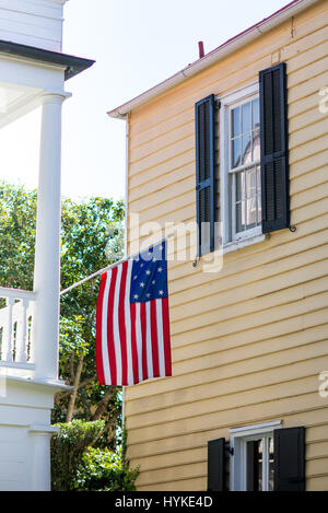 Un drapeau américain est suspendu off white porche coloniale adjacent à une maison jaune à Charleston, Caroline du Sud pendant les mois d'été. Banque D'Images