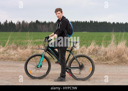 Young man on mountain bike se tient sur chemin, sur fond de verte prairie et forêt. Banque D'Images