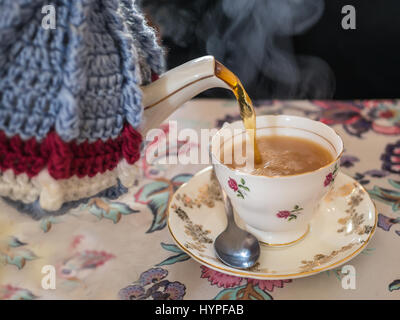 On verse le thé d'un vintage Bone China Tea pot avec laine tricoté avec couvercle confortable dans une jolie tasse à thé avec soucoupe. Le tout sur un modèle style vintage Banque D'Images