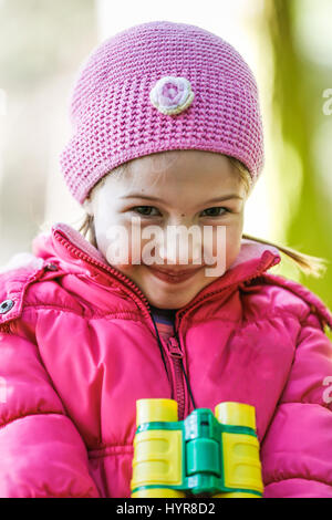 Portrait d'une petite fille en rose avec des jumelles autour du cou, explorer la nature, être espiègle et timide pour l'appareil photo. Style de vie en plein air actif c Banque D'Images