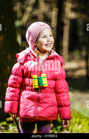 Petite fille aux jumelles, enjoying sun au cours de promenade dans la forêt, explorer la nature, être espiègle et timide pour appareil photo. Style de vie en plein air actif con Banque D'Images