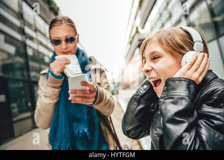 L'écoute de la musique sur le casque fille avec mère debout à côté d'alimentation et d'utilisation de smart phone Banque D'Images