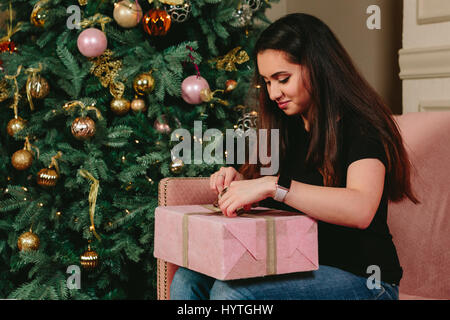 Belle jeune femme brune sur un canapé déballe le cadeau près de l'arbre de Noël. Studio portrait horizontal. Banque D'Images