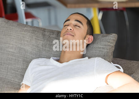 Jeune homme noir en blanc T-shirt détente sur canapé gris à l'intérieur allongé sur grand coussin avec ses yeux fermés Banque D'Images